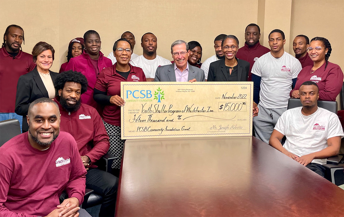 PCSB Community Foundation Chair Joe Roberto presents an oversized grant check to the Youth Shelter Program of Westchester Inc, and is joined by a large group of nonprofit staff from that organization who huddle around the check presentation.